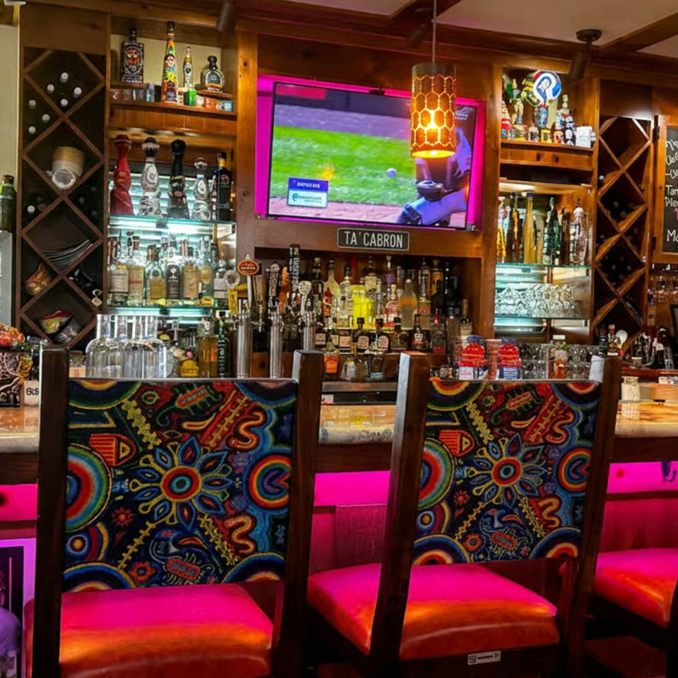 Colorful restaurant bar with liquor bottles, beer taps, television, patterned chairs, and vibrant pink lighting.
